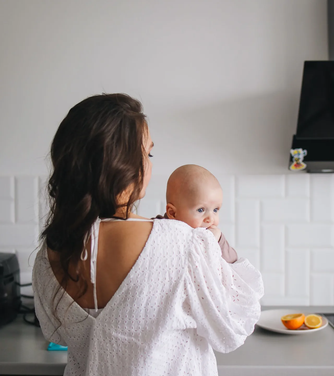 A mother in a white dress holding her baby in a bright, light-filled kitchen.
